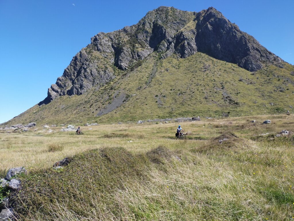 Two riders crossing open tussock beneath a steep coastal hill